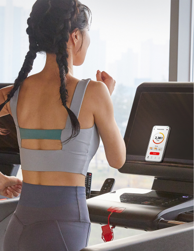 A woman is running on a treadmill at the gym. The screen of a smartphone placed on the treadmill is clearly visible.