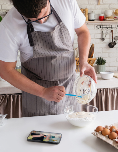 A man is cooking in the kitchen, wearing an apron, with a clear view of the smartphone display next to him.