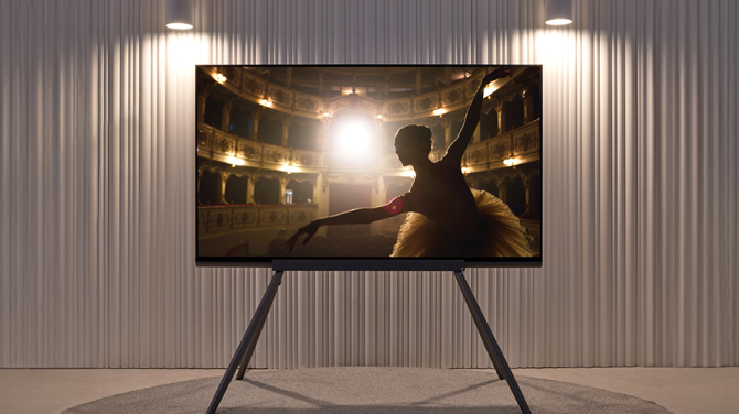 The transparent OLED panel is positioned in a well-lit living room setting with white curtains. The screen shows the graceful gestures of a ballerina performing in a theater.