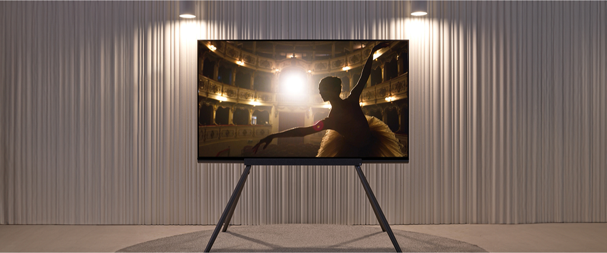 The transparent OLED panel is positioned in a well-lit living room setting with white curtains. The screen shows the graceful gestures of a ballerina performing in a theater.